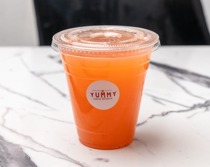A plastic cup of orange juice with a "Yummy Italian Taste" sticker sits on a marble surface.