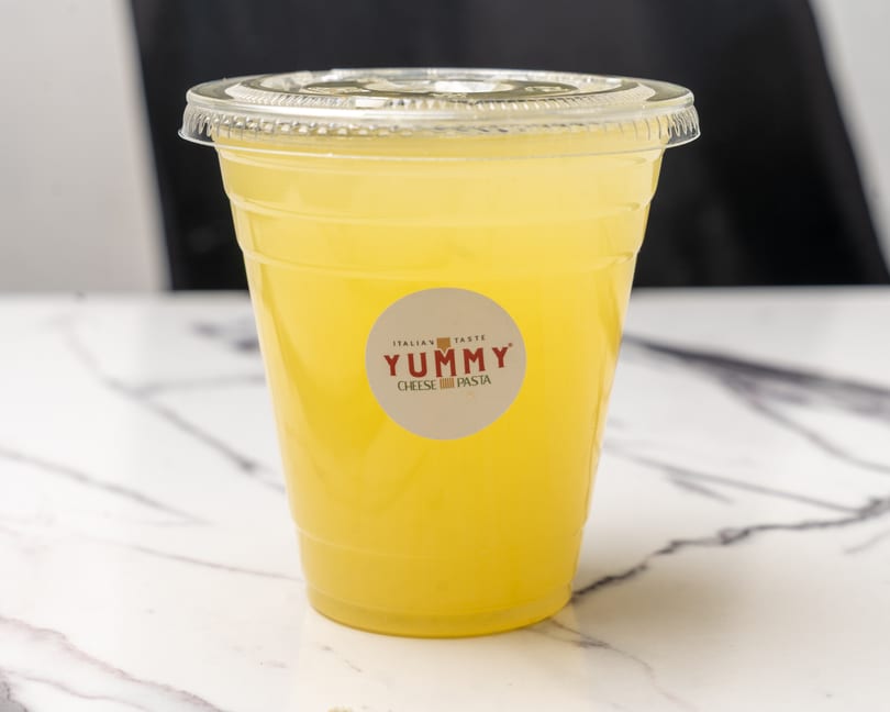 A clear plastic cup of yellow lemonade with a Yummy Cheese Pasta logo on a marble surface.