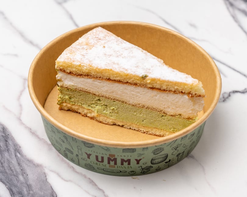 A slice of layered cake with green and white filling, topped with powdered sugar, in a Yummy branded container.
